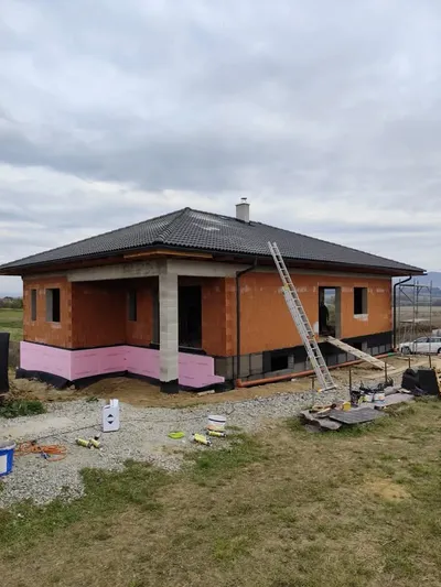 Dokončený prízemný bungalov s valbovou strechou z tmavej betónovej škridly a tehlovými stenami s izolačným pásom pri základoch