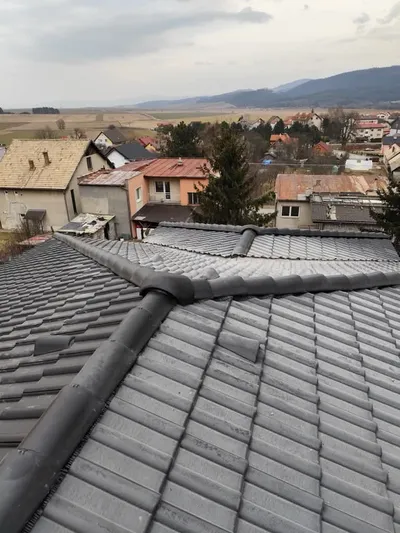 Pohľad zblízka na hrebeň a nároží strechy pokrytej tmavou betónovou škridlou s hrebenáčmi, v pozadí dedina a kopce